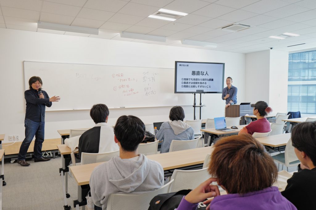 DBM学科の講義中、学科長の金谷と曽山さんが会話しているシーン